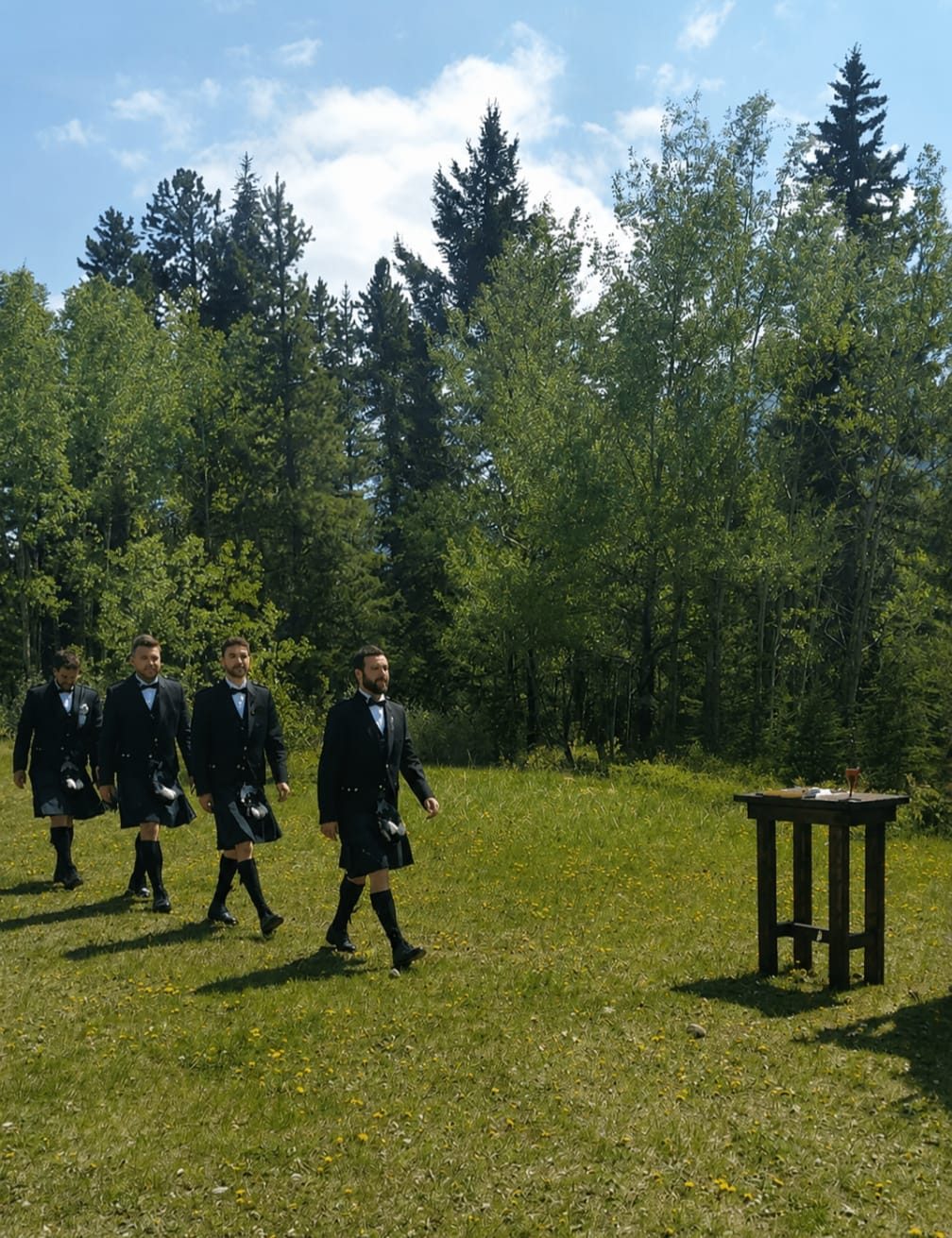 Groomsmen in full Highland dress processing across a lawn to a ceremony table at a real wedding